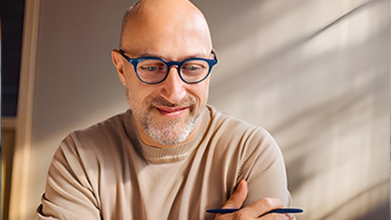 A man wearing glasses thinking and smiling with a pen in hand.